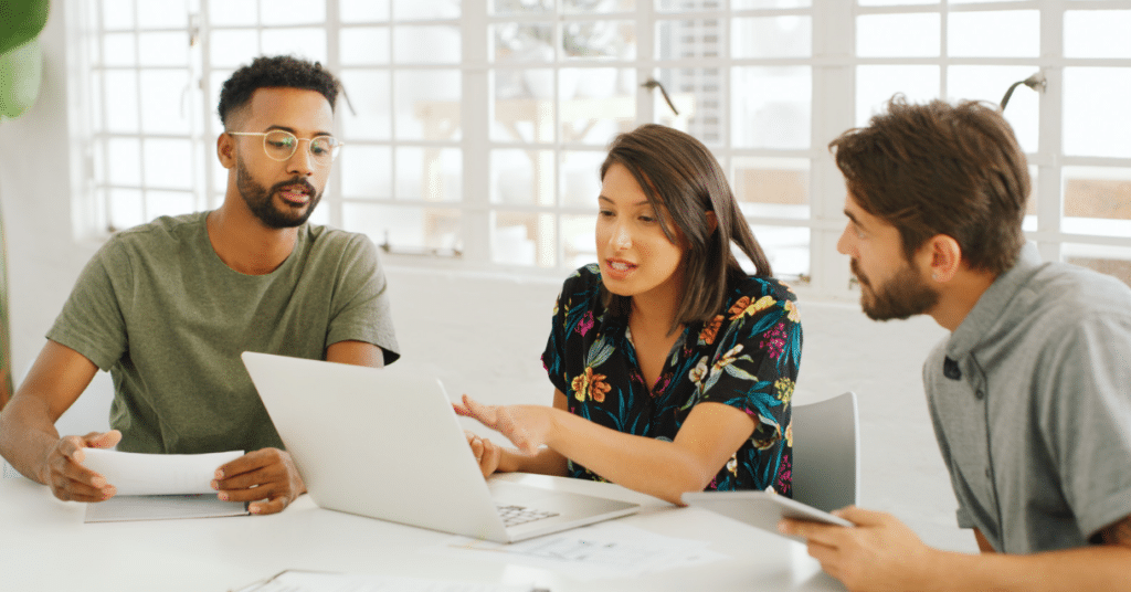 Three casually-dressed office workers chatting at a table and looking at a laptop screen.