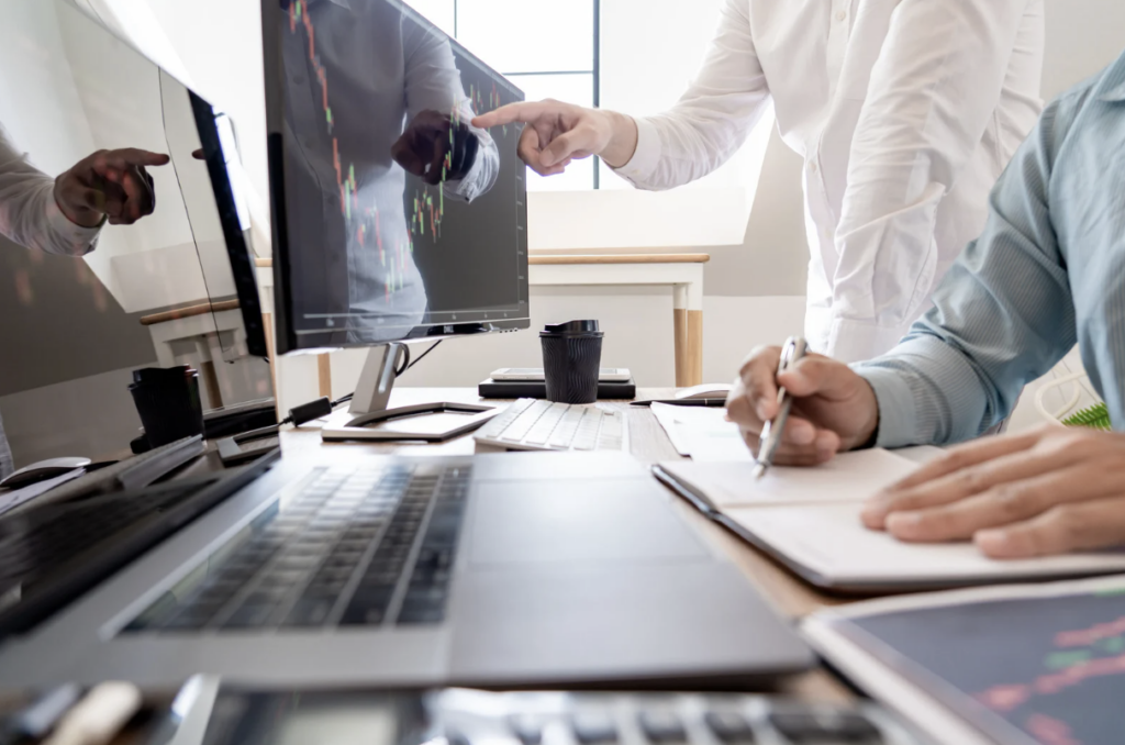 office worker sat at desk taking notes and being advised by co-worker pointing at computer screen