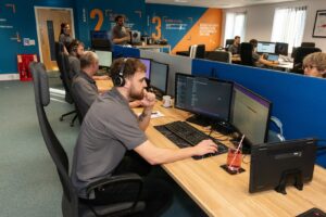 Service desk engineer wearing a headset at a multi-monitor workstation, with colleagues working at desks behind him in the Your IT Department office