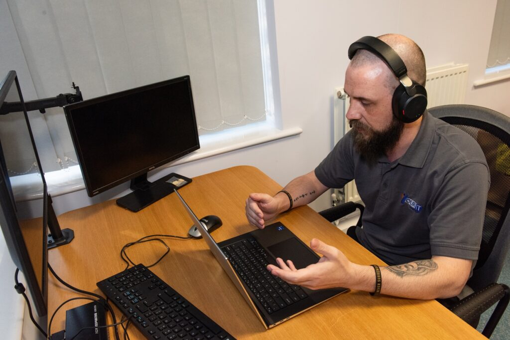 An IT support technician wearing a "Your IT" branded polo shirt and headphones speaks during a video call at a wooden desk with a laptop, external keyboard, mouse, and two monitors, demonstrating how remote support is delivered in a professional work environment.